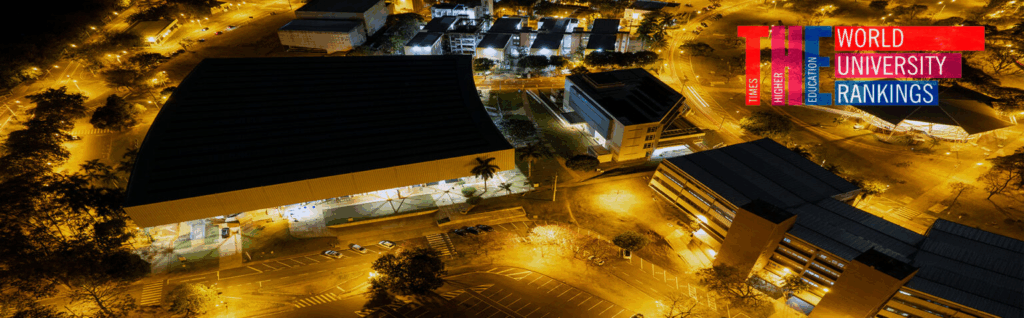 Uma fotografia aérea noturna mostra um campus universitário iluminado, com vários edifícios e áreas verdes, juntamente com a logomarca "World University Rankings".
A composição da imagem mostra uma perspectiva aérea da universidade, com edifícios, estradas e áreas ajardinadas. A iluminação artificial, provavelmente de postes de luz e holofotes, lança um brilho quente em toda a cena, com contraste entre as áreas escuras e iluminadas. A disposição dos edifícios e espaços cria uma sensação de ordem e organização. No canto superior direito, há um logotipo "World University Rankings" que adiciona contexto ao tema.
Os elementos principais são os edifícios do campus, com suas arquiteturas distintas e iluminação, e a vegetação espalhada entre eles. A iluminação artificial define a forma e a textura dos edifícios. A presença da logo "World University Rankings" é um ponto focal que indica a relevância da universidade no cenário global de educação superior.
A fotografia utiliza uma técnica de fotografia aérea, possivelmente tirada de um drone ou de um ponto de vista elevado, que permite uma ampla visão do campus universitário. O estilo de execução é realista, com atenção aos detalhes da arquitetura e do ambiente. As escolhas criativas incluem o uso da iluminação noturna para destacar os edifícios e criar uma atmosfera de atividade e conhecimento.
O cenário é um campus universitário noturno, com a iluminação artificial criando um contraste notável com o céu escuro. A atmosfera é calma e focada, sugerindo um ambiente acadêmico. A iluminação quente, combinada com a arquitetura moderna dos edifícios, cria uma sensação de progresso e inovação. A presença da logomarca "World University Rankings" reforça a ideia de uma instituição de ensino de renome mundial.