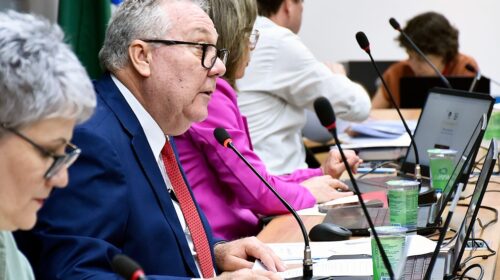 Fotografia que mostra pessoas em mesa falando em microfones. Em primeiro plano, ao centro, há um homem branco, grisalho, usando óculos de grau e vestindo roupas sociais. À frente do homem, há uma mulher branca, grisalha, usando óculos de grau e vestindo roupas sociais. Ao fundo há mais pessoas. O ambiente é fechado e bem iluminado. Fim da descrição.