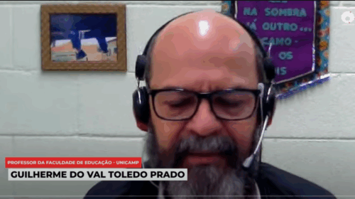 Captura de tela de computador que mostra parte de vídeo no YouTube. Há um homem branco, grisalho e usando óculos de grau e fones de ouvido ao centro. Ao fundo, há uma parede clara com enfeites. Há duas caixas de texto alinhadas à lateral esquerda, com os textos "Professor da Faculdade de Educação - Unicamp" e "Guilherme do Val Toledo Prado". Fim da descrição.