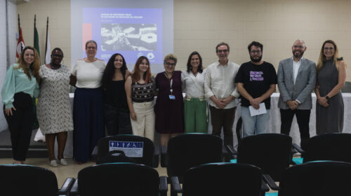 Fotografia que mostra um grupo de 11 pessoas sorrindo e posando para a câmera. O ambiente é uma sala fechada e bem iluminada e há cadeiras, mesa, bandeiras no ambiente. As pessoas vestem roupas formais. Fim da descrição.