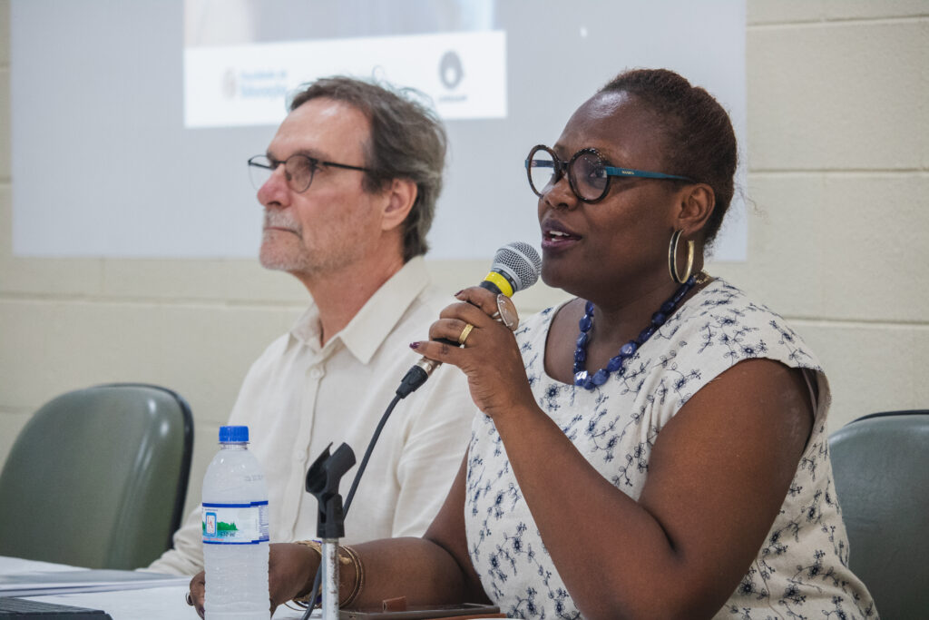 Fotografia que mostra duas pessoas sentadas à mesa em evento. À frente na fotografia, há uma mulher segurando um microfone; ela é negra, tem cabelo castanho preso, veste blusa branca estampada e usa óculos de grau com armação azul e colar azul. Ao lado da mulher, há um homem olhando para frente. O homem é branco, tem cabelo e barba grisalhos, veste camisa branca e usa óculos de grau com armação preta. Ao fundo, há uma parede branca. Fim da descrição.