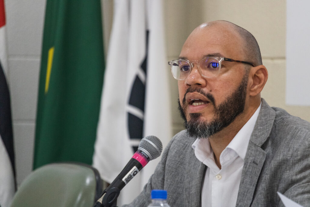 Fotografia que mostra um homem sentado falando no microfone e olhando para frente. O homem tem tom de pele marrom claro e barba grisalha e bem definida; veste camisa branca e terno cinza e usa óculos de grau com armação transparente. Ao fundo, há bandeiras. Fim da descrição.