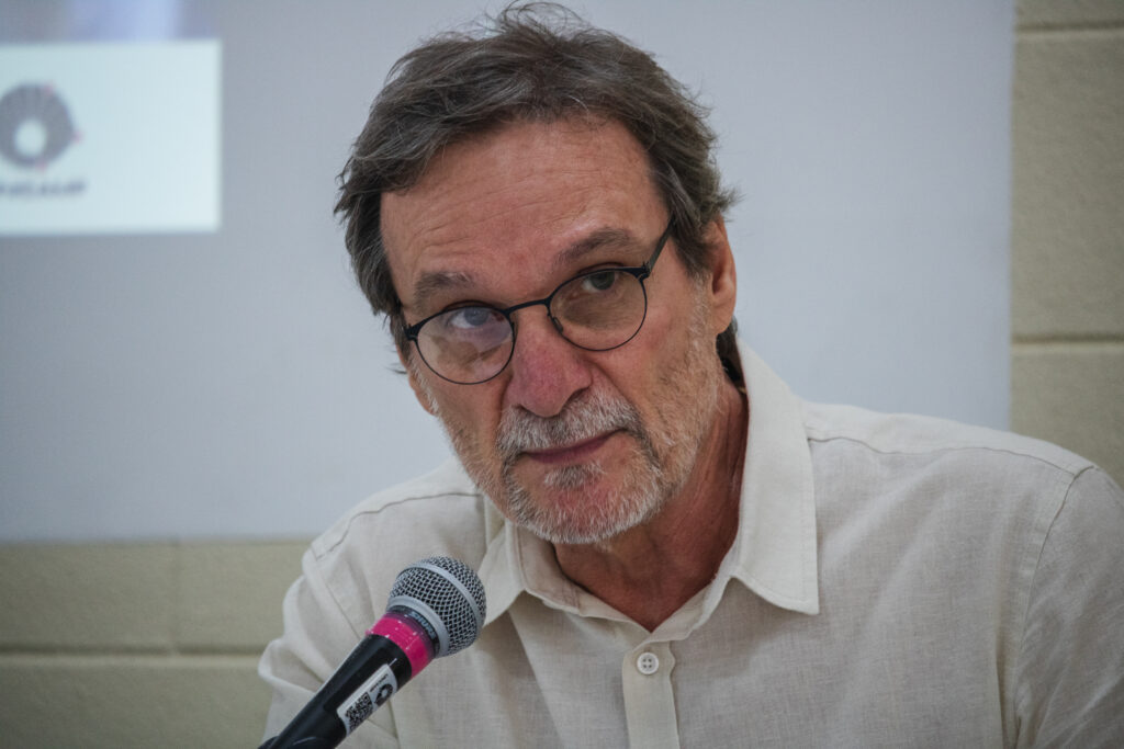 Fotografia que mostra homem olhando para frente. O homem é branco, tem cabelo e barba grisalhos, veste camisa branca e usa óculos de grau com armação preta. À sua frente, há um microfone. Ao fundo, há uma parede branca. Fim da descrição. 