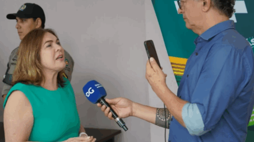 Fotografia que mostra uma mulher sendo entrevistada por um homem. A mulher é branca, tem cabelo curto e loiro e usa blusa verde e o homem é branco, usa camisa jeans e segura um microfone azul e um celular. O ambiente é bem iluminado. Fim da descrição.