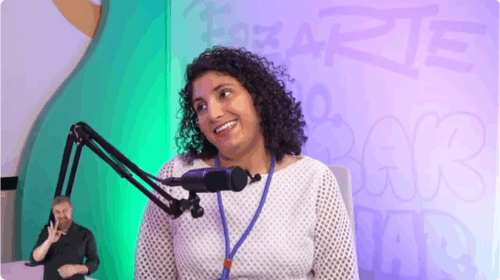 Captura de tela que mostra mulher em estúdio de podcast. A mulher é parda, tem cabelo cacheado, curto e castanho, veste blusa branca e sorri. O fundo é colorido em tons de roxo e verde. Há um microfone à sua frente. Fim da descrição.