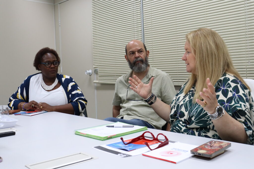 Fotografia que mostra três pessoas conversando em sala. As três pessoas estão sentadas à mesa, onde há cadernos, papéis, canetas e celulares. Da esquerda para a direita: há uma mulher negra com cabelo curto, crespo e castanho vestindo blusa branca, casaco estampado e colar grande branco e usando óculos de grau; há um homem branco com barba grande e grisalha vestindo camisa bege e há uma mulher branca com cabelo curto e loiro vestindo blusa azul e branca estampada e gesticulando com as mãos. O ambiente é bem iluminado e tem paredes claras. Fim da descrição.