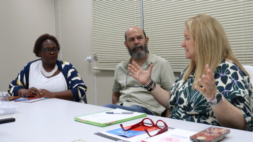 Fotografia que mostra três pessoas conversando em sala. As três pessoas estão sentadas à mesa, onde há cadernos, papéis, canetas e celulares. Da esquerda para a direita: há uma mulher negra com cabelo curto, crespo e castanho vestindo blusa branca, casaco estampado e colar grande branco e usando óculos de grau; há um homem branco com barba grande e grisalha vestindo camisa bege e há uma mulher branca com cabelo curto e loiro vestindo blusa azul e branca estampada e gesticulando com as mãos. O ambiente é bem iluminado e tem paredes claras. Fim da descrição.