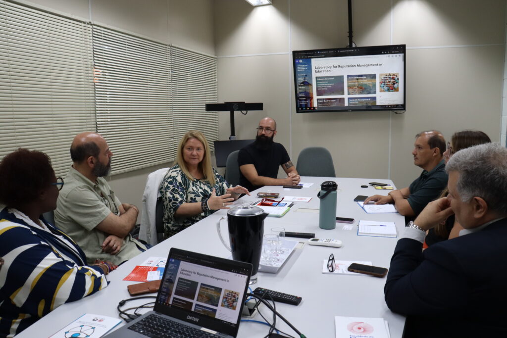 Fotografia que mostra pessoas conversando em sala. O ambiente é fechado, bem iluminado, há paredes claras e uma grande mesa ao centro da sala com papéis, cadernos, garrafas, copos, notebook, óculos e outros itens de escritório. As pessoas observam uma mulher falar. Fim da descrição.