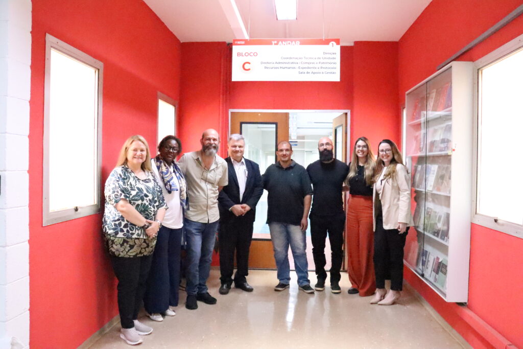 Fotografia que mostra grupo de oito pessoas, quatro mulheres e quatro homens, sorrindo e posando para a câmera uma ao lado da outra. O ambiente é fechado, bem iluminado e tem paredes vermelhas. Há uma vitrine na parede com livros e uma placa pendurada no teto com a indicação de que o grupo está no "Bloco C". Fim da descrição.