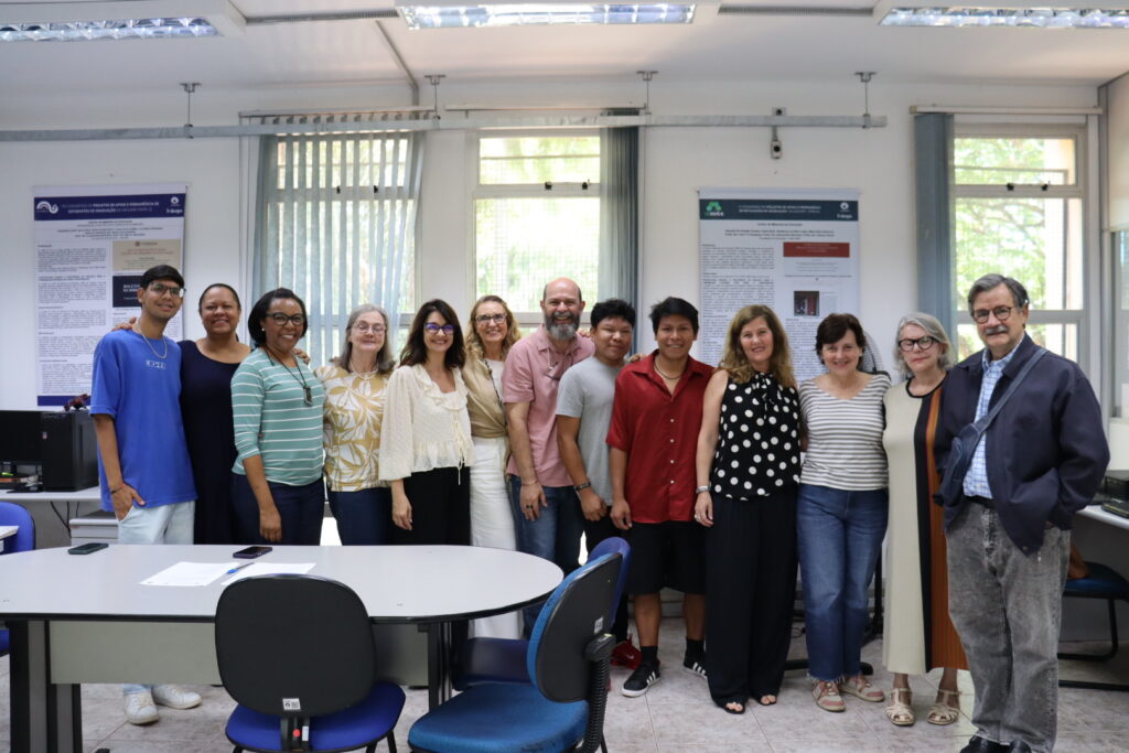 Fotografia que mostra grupo de treze pessoas, uma ao lado da outra, sorrindo e olhando para a câmera. O ambiente é uma sala fechada e bem iluminada, a parede é branca e há grandes janelas com cortinas azuis. Na sala, há mesas, cadeiras, banners, papéis e aparelhos eletrônicos. Fim da descrição.