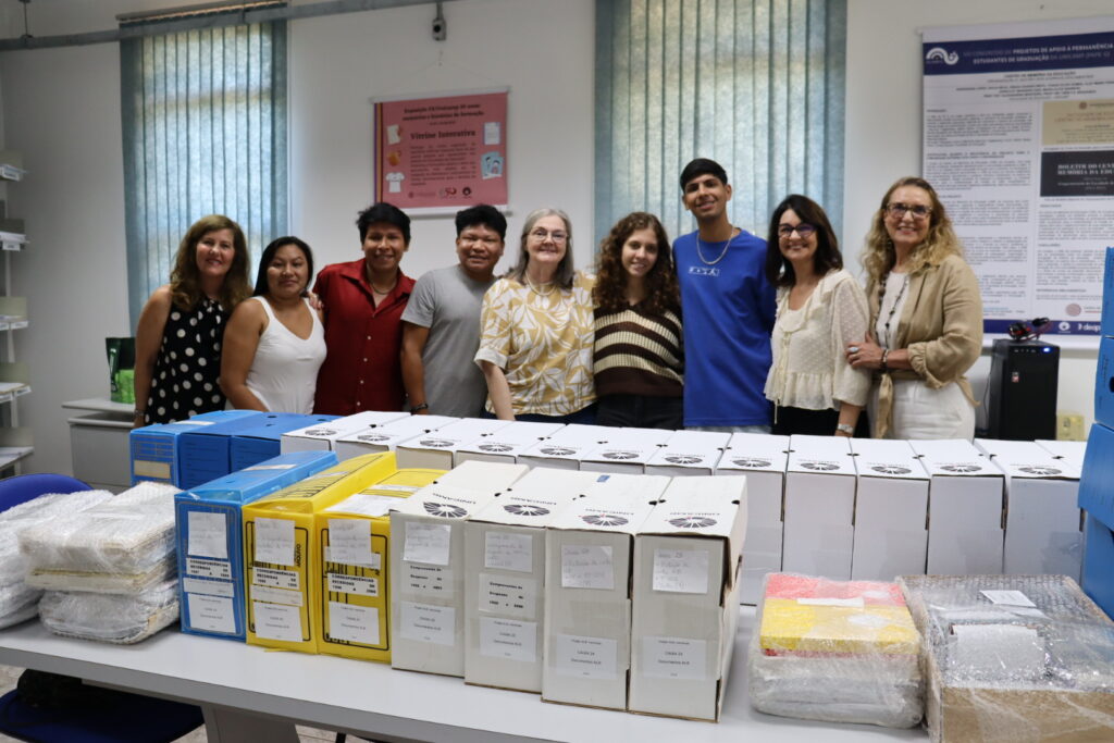 Fotografia que mostra grupo de nove pessoas, uma ao lado da outra, sorrindo e olhando para a câmera. O ambiente é uma sala fechada e bem iluminada, a parede é branca com cortinas azuis. À frente das pessoas, há uma mesa com papéis cadernos envoltos por plástico bolha e aproximadamente vinte caixas de papelão. Fim da descrição.