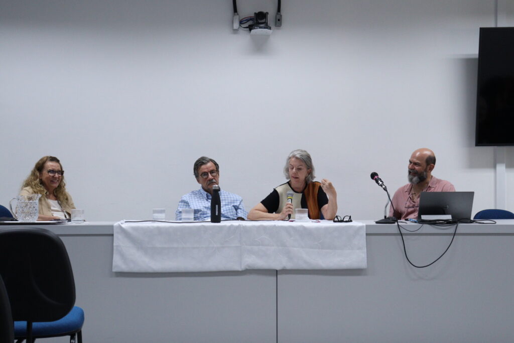 Fotografia que mostra quatro pessoas sentadas à mesa em uma sala. O ambiente é fechado e bem iluminado, a parede é branca e a mesa é comprida e branca, há jarra e copos de água, microfones, notebook, televisão e cadeiras na sala. Da esquerda para a direita: mulher branca com cabelo loiro, comprido e ondulado, veste blusa branca e blazer bege e usa óculos de grau; homem branco com cabelo grisalho, curto e liso, veste camisa azul quadriculada e usa óculos de grau; mulher branca com cabelo grisalho, curto e liso, veste blusa preta, marrom e branca e segura um microfone e homem branco calvo com barba grande e grisalha, veste camisa rosa. Fim da descrição.