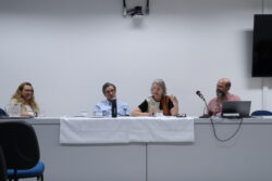 Fotografia que mostra quatro pessoas sentadas à mesa em uma sala. O ambiente é fechado e bem iluminado, a parede é branca e a mesa é comprida e branca, há jarra e copos de água, microfones, notebook, televisão e cadeiras na sala. Da esquerda para a direita: mulher branca com cabelo loiro, comprido e ondulado, veste blusa branca e blazer bege e usa óculos de grau; homem branco com cabelo grisalho, curto e liso, veste camisa azul quadriculada e usa óculos de grau; mulher branca com cabelo grisalho, curto e liso, veste blusa preta, marrom e branca e segura um microfone e homem branco calvo com barba grande e grisalha, veste camisa rosa. Fim da descrição.