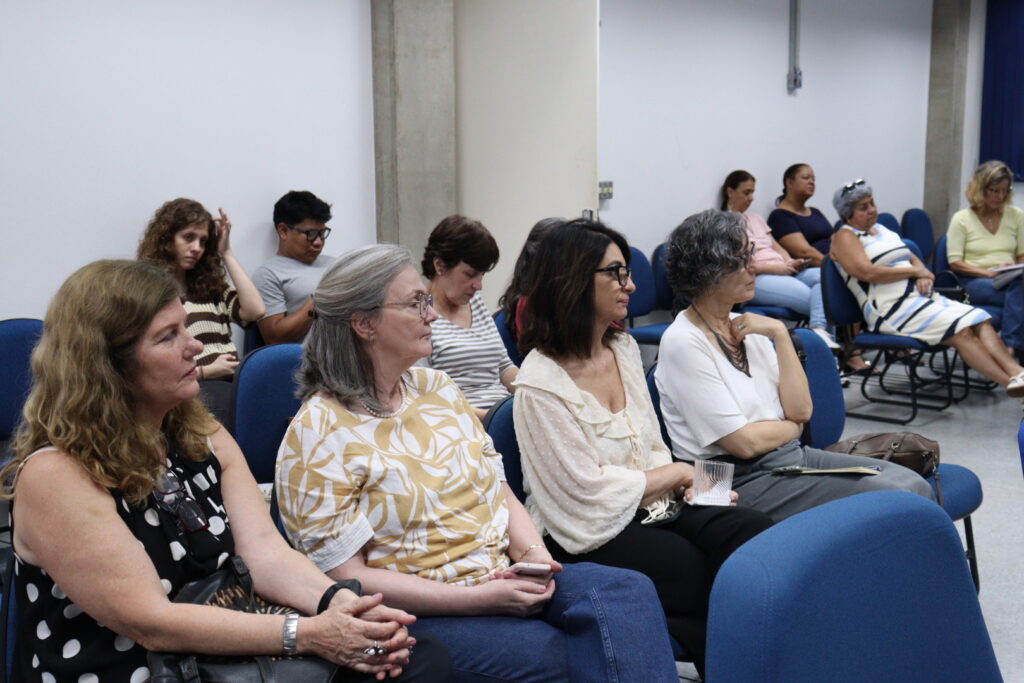 Fotografia que mostra público assistindo a algo. O ambiente é uma sala bem iluminada com paredes brancas, cortinas azuis e cadeiras azuis enfileiradas. As pessoas olham para frente com expressões de concentração. Fim da descrição.