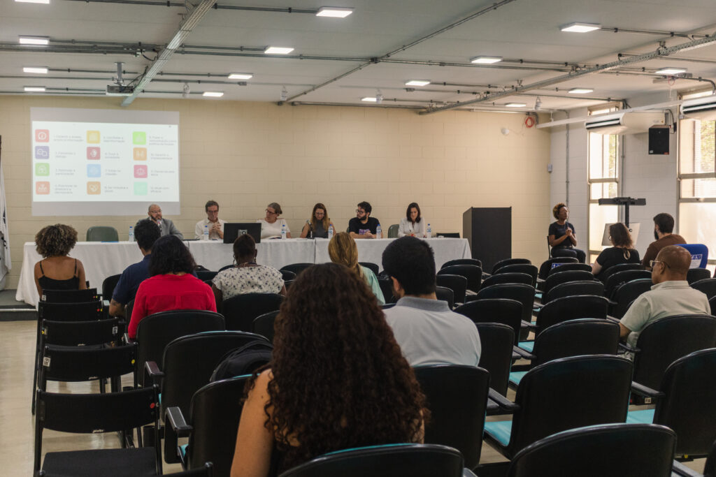 Fotografia que mostra mesa temática em evento institucional. O ambiente é fechado e bem iluminado, as paredes e o teto são claros e há cadeiras enfileiradas, onde há pessoas sentadas. À frente das pessoas sentadas, está a mesa temática, com seis pessoas sentadas. Fim da descrição.