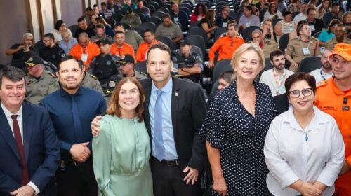 Fotografia que mostra um grupo de seis pessoas olhando e posando para a câmera. As pessoas vestem roupas formais. Ao redor e ao fundo, há mais pessoas. O ambiente se assemelha a um salão, com cadeiras enfileiradas. Fim da descrição.