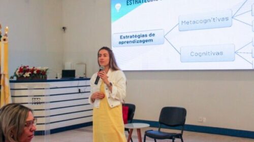 Fotografia que mostra professora em sala de aula. A sala está parcialmente iluminada e há uma projeção de imagem na parede, atrás da professora. Há mesa e cadeiras na sala. A professora é branca, tem cabelo liso e loiro e usa vestido amarelo e jaqueta branca, ela segura um microfone. Fim da descrição.