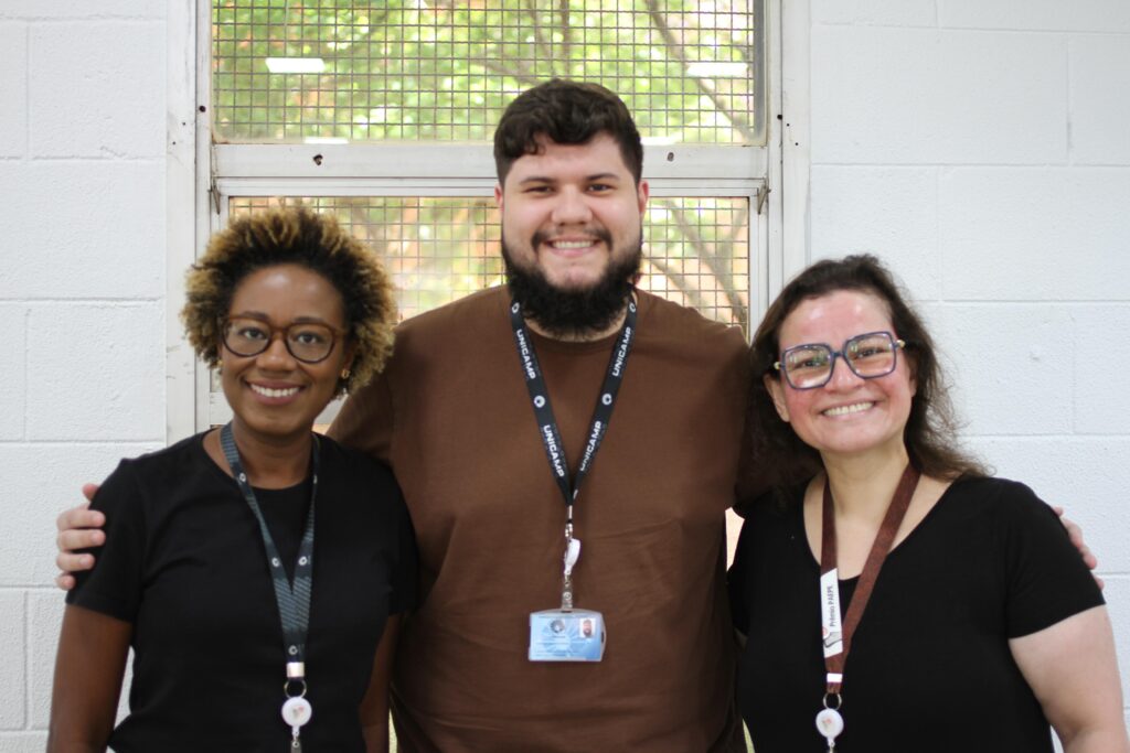 Fotografia que mostra três pessoas, uma ao lado da outra, sorrindo e olhando para a câmera. Da esquerda para a direita: há uma mulher com pele marrom escuro e cabelo loiro, crespo e curto e vestindo camiseta preta e usando óculos de grau com armação marrom e cordão de crachá preto; há um homem com pele bege claro e cabelo castanho, liso e curto e castanho e barba da mesma cor vestindo camiseta marrom e usando cordão de crachá preto com crachá azul pendurado e há uma mulher o pele bege rosado e cabelo castanho, liso e curto vestindo blusa preta e usando óculos de grau com armação azul e cordão de crachá marrom. Ao fundo, há uma parede clara com janela grande. Fim da descrição.
