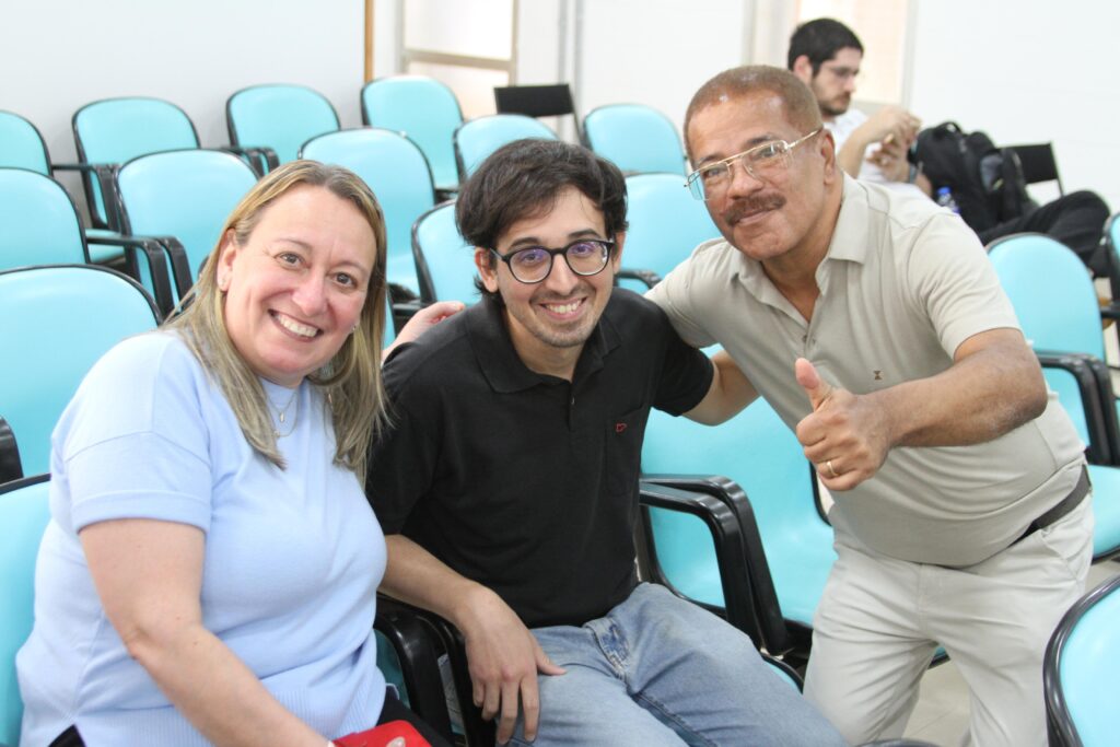 Fotografia que mostra três pessoas olhando e sorrindo para a câmera em salão com cadeiras azuis enfileiradas. Da esquerda para a direita: há uma mulher com pele bege rosado e cabelo liso e loiro vestindo blusa azul claro; há um homem com pele bege claro e cabelo liso e castanho vestindo camisa preta e calça jeans e usando óculos de grau de armação preta e há um homem com pele marrom claro e cabelo e bigode loiro escuro vestindo camisa bege e calça branca. Fim da descrição.

