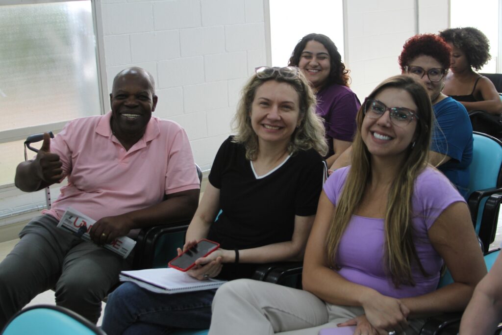 Fotografia que mostra três pessoas sentadas em público de evento. Da esquerda para a direita: homem negro, careca, vestindo blusa rosa e calça cinza; mulher branca, com cabelo loiro, liso e curto, vestindo blusa preta e calça jeans e mulher branca, com cabelo loiro, liso e comprido, vestindo blusa lilás e calça bege e usando óculos de grau. Ao fundo, há mais pessoas sentadas. O ambiente é fechado e bem iluminado. Fim da descrição.