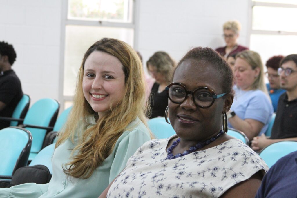Fotografia que mostra duas mulheres em público de evento. Elas sorriem para a câmera. A mulher da esquerda é branca, tem cabelo loiro, liso e comprido e usa camisa verde. A mulher da esquerda é negra, tem cabelo castanho, crespo e preso e usa blusa branca estampada, óculos de grau com armação colorida, colar azul grande e brincos de argola grandes. Ao fundo, há mais pessoas compondo o público do evento. O ambiente é fechado e há janelas por onde entra iluminação solar. Fim da descrição.
