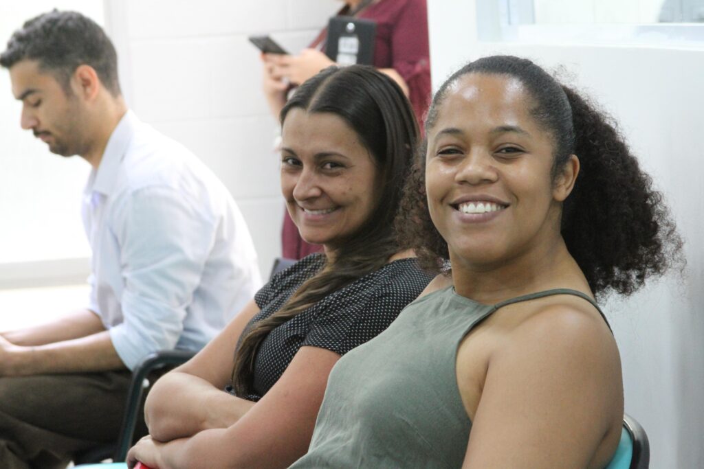 Fotografia que mostra duas mulheres em público de evento. Elas sorriem para a câmera. A mulher da esquerda é parda, tem cabelo castanho, liso e comprido e veste blusa preta de bolinhas brancas. A mulher da esquerda é negra, tem cabelo castanho, cacheado e preso e usa blusa verde. Ao fundo, há mais pessoas compondo o público do evento. O ambiente é fechado e bem iluminado. Fim da descrição.