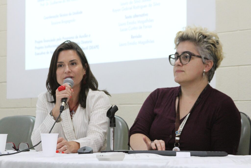 Fotografia que mostra duas mulheres sentadas à mesa falando em evento. À esquerda, há uma mulher branca, com cabelo liso, castanho e tamanho médio, vestindo blazer branco com listras pretas e falando no microfone. À direita, há uma mulher branca, com cabelo liso, loiro e curto, vestindo blusa roxa e usando óculos de grau com armação preta. Ao fundo, há uma parede branca. Fim da descrição. 