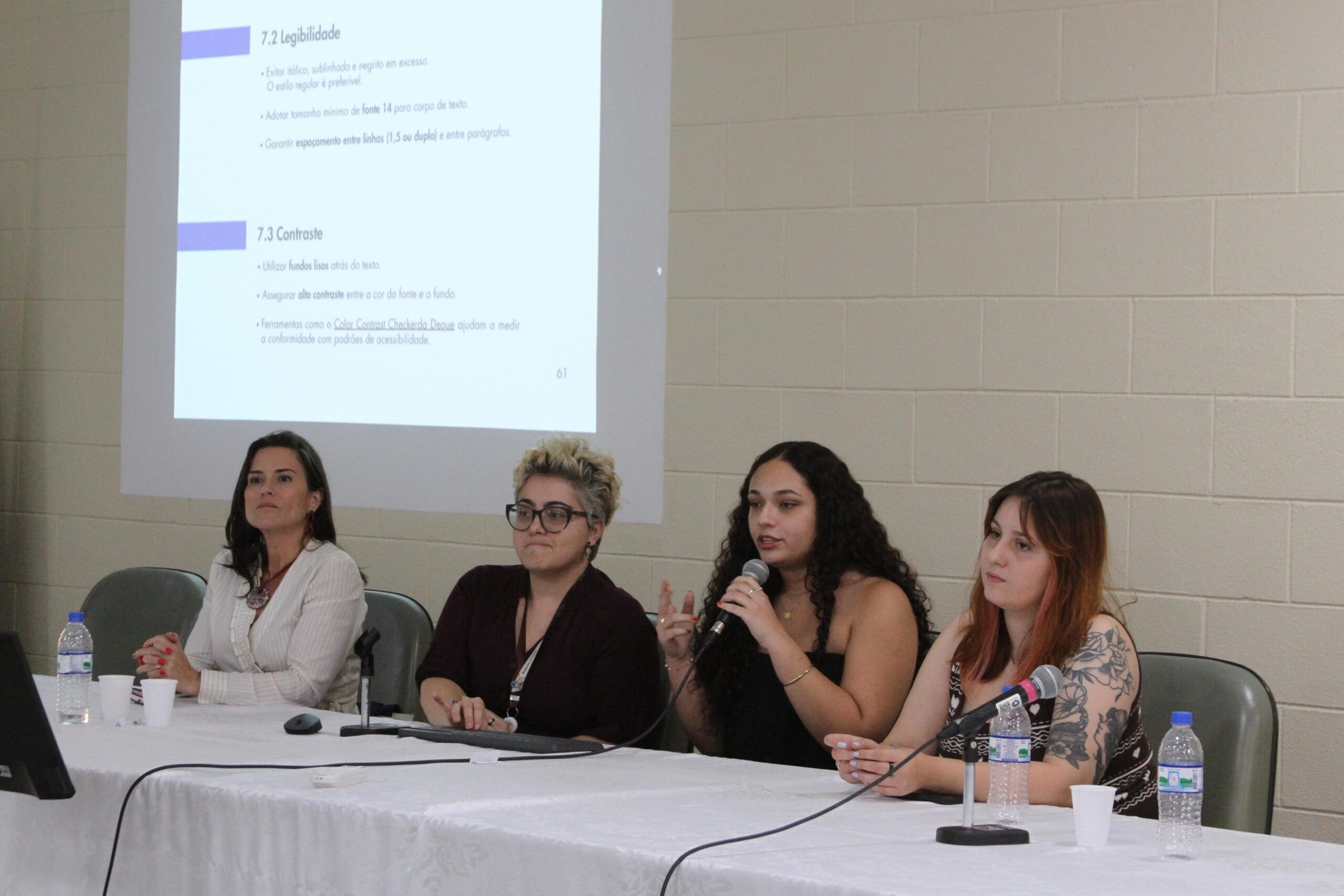 Fotografia que mostra quatro mulheres sentadas à mesa falando em evento. Da esquerda para a direita: mulher branca com cabelo castanho liso vestindo blazer branco; mulher branca com cabelo loiro liso vestindo blusa roxa; mulher parda com cabelo castanho cacheado vestindo blusa preta e segurando um microfone e mulher branca com cabelo ruivo liso. Na mesa, há microfones e copos de água. Ao fundo, há uma parede branca. Fim da descrição.