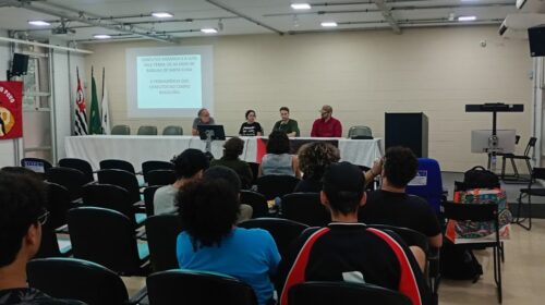 Fotografia que mostra evento em salão. Há pessoas na plateia e à frente há uma mesa temática. Fim da descrição.