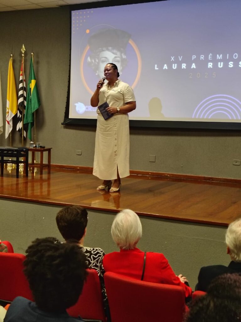 Fotografia que mostra mulher falando em palco de auditório. A mulher é negra, tem cabelo trançado preto e veste camisa e saia brancas. O ambiente é fechado, as paredes são escuras, o palco é de madeira, há cadeiras vermelhas enfileiradas à frente do palco. No palco, há uma projeção de imagem com a escrita "XV Prêmio Laura Russo 2025". Fim da descrição.