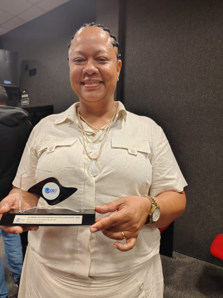 Fotografia que mostra mulher segurando prêmio. A mulher é negra, tem cabelo trançado preto, veste camisa e saia brancas e usa acessórios dourados. O ambiente é fechado e bem iluminado e as paredes são escuras. Fim da descrição.