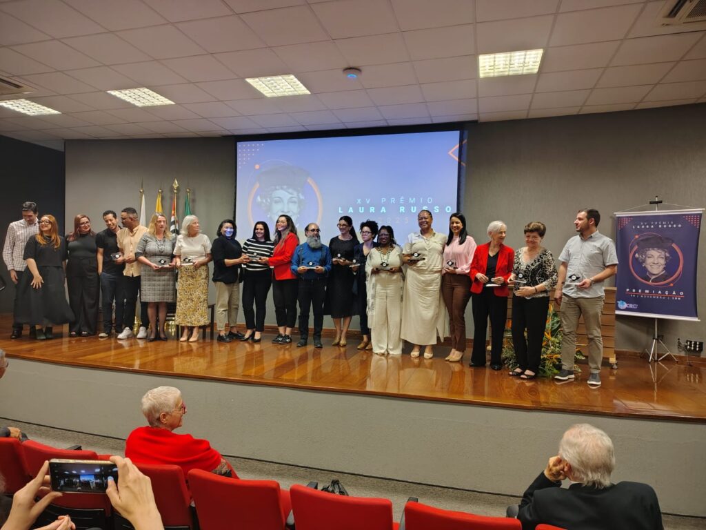 Fotografia que mostra um grupo de pessoas em um palco de auditório. As pessoas vestem roupas formais e seguram estatuetas de prêmio. O ambiente é fechado, as paredes são escuras, o palco é de madeira, há cadeiras vermelhas enfileiradas à frente do palco. No palco, há um banner e uma projeção de imagem com a escrita "XV Prêmio Laura Russo". Fim da descrição.
