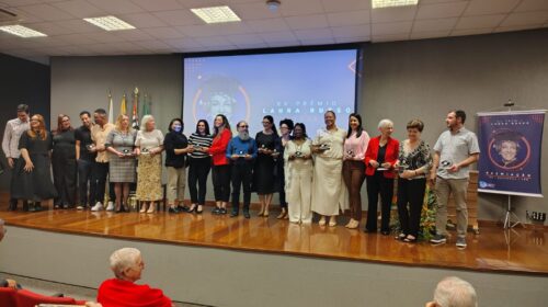 Fotografia que mostra um grupo de pessoas em um palco de auditório. As pessoas vestem roupas formais e seguram estatuetas de prêmio. O ambiente é fechado, as paredes são escuras, o palco é de madeira, há cadeiras vermelhas enfileiradas à frente do palco. No palco, há um banner e uma projeção de imagem com a escrita "XV Prêmio Laura Russo". Fim da descrição.