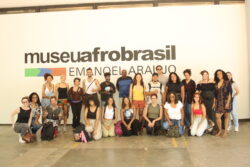 Fotografia que mostra um grupo de pessoas posando para a câmera à frente de uma parede com a escrita "museu afro brasil". Fim da descrição.