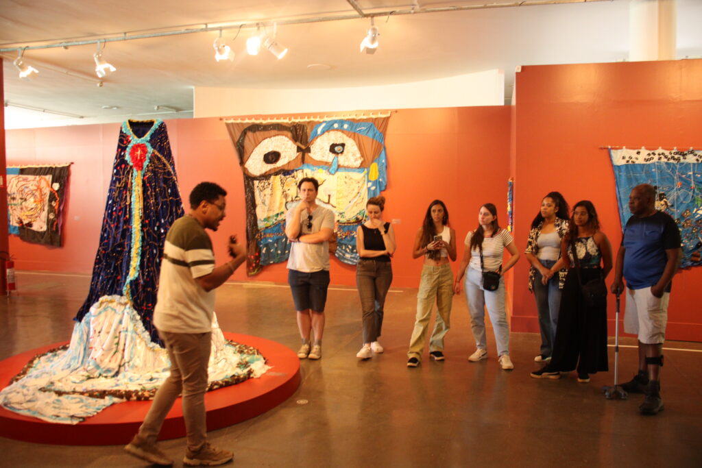 Fotografia que mostra grupo de pessoas em Museu. O ambiente é fechado com iluminação amarela, as paredes são laranja e há obras de arte nas paredes e no meio do ambiente. Fim da descrição.