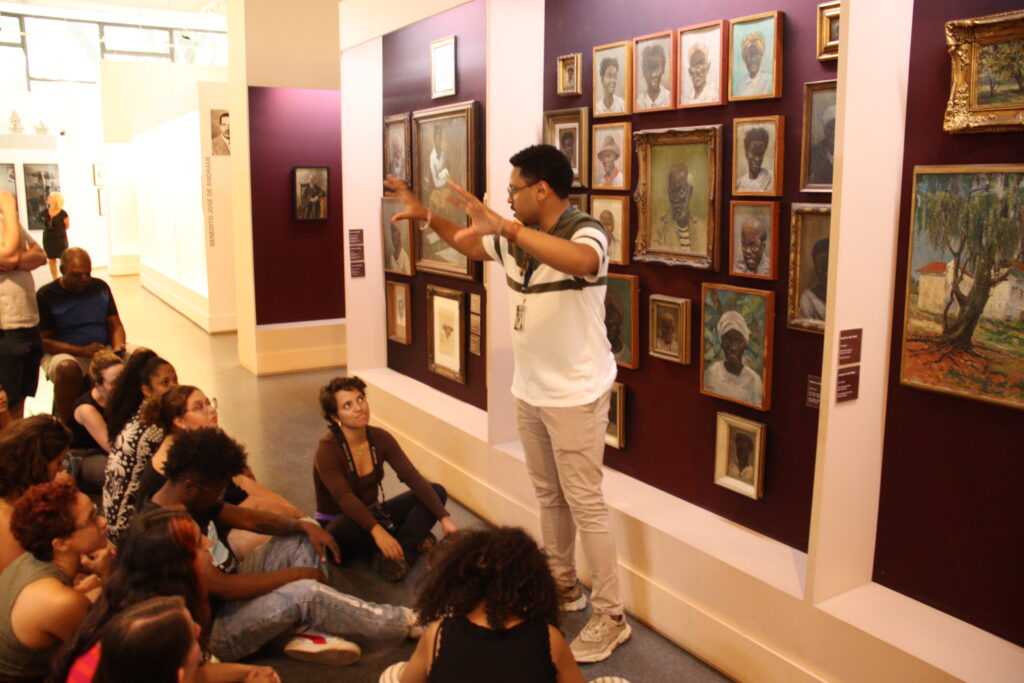 Fotografia que mostra um grupo de pessoas em um museu. O ambiente é fechado, as paredes são brancas e roxas e a iluminação é amarela. Há quadros pendurados nas paredes. Fim da descrição.