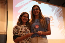 Fotografia que mostra duas mulheres segurando um prêmio. O ambiente é fechado e parcialmente iluminado. A mulher da esquerda é branca, tem cabelo loiro, liso e curto e veste um vestido estampado e colorido. A mulher da direita é branca, tem cabelo loiro, liso e longo e veste uma blusa azul e uma calça branca. As duas sorriem e olham para a câmera. Fim da descrição.