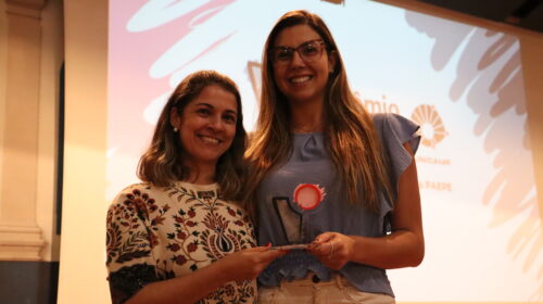 Fotografia que mostra duas mulheres segurando um prêmio. O ambiente é fechado e parcialmente iluminado. A mulher da esquerda é branca, tem cabelo loiro, liso e curto e veste um vestido estampado e colorido. A mulher da direita é branca, tem cabelo loiro, liso e longo e veste uma blusa azul e uma calça branca. As duas sorriem e olham para a câmera. Fim da descrição.