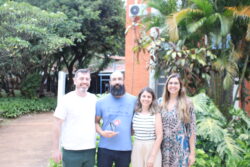 Fotografia que mostra quatro pessoas em pé, uma ao lado da outra, sorrindo e olhando para a câmera em ambiente externo. Da esquerda para a direita: homem branco com cabelo e barba grisalhos veste camiseta branca e calça jeans; homem branco com barba grande e preta veste camiseta azul e calça jeans e segura um prêmio; mulher branca com cabelo liso, curto e loiro veste blusa branca listrada e calça bege e mulher branca com cabelo liso, longo e loiro veste vestido estampado em tons de preto, branco e azul. O ambiente é aberto e bem iluminado, há árvores e plantas e um prédio institucional ao fundo. Fim da descrição.