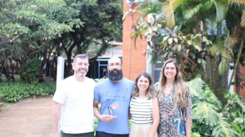 Fotografia que mostra quatro pessoas em pé, uma ao lado da outra, sorrindo e olhando para a câmera em ambiente externo. Da esquerda para a direita: homem branco com cabelo e barba grisalhos veste camiseta branca e calça jeans; homem branco com barba grande e preta veste camiseta azul e calça jeans e segura um prêmio; mulher branca com cabelo liso, curto e loiro veste blusa branca listrada e calça bege e mulher branca com cabelo liso, longo e loiro veste vestido estampado em tons de preto, branco e azul. O ambiente é aberto e bem iluminado, há árvores e plantas e um prédio institucional ao fundo. Fim da descrição.