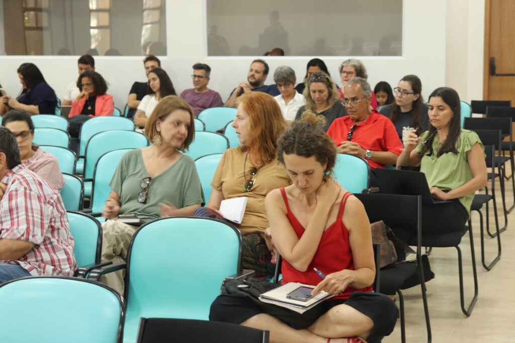Fotografia que mostra a plateia de um evento. O ambiente é fechado e bem iluminado, as paredes são claras e há cadeiras azuis enfileiradas. As pessoas estão sentadas nas cadeiras e olham para frente, algumas conversam entre si, algumas seguram papéis e canetas. Fim da descrição.