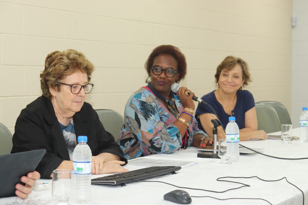 Fotografia que mostra três mulheres sentadas em uma mesa de evento. Da esquerda para a direita: mulher branca, cabelo loiro, ondulado e curto, veste blusa azul estampada e blazer preto e usa óculos de grau; mulher negra, cabelo castanho, crespo e curto, veste blusa estampada em tons de azul e rosa, usa braceletes, colar e brincos grandes e óculos de grau e segura um microfone; mulher branca, cabelo loiro, liso e curto, veste blusa azul e usa brincos dourados. O fundo é uma parede clara e em cima da mesa há teclado, mouse, garrafas de água e notebook. Fim da descrição.