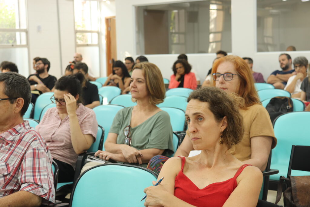 Fotografia que mostra a plateia de um evento. O ambiente é fechado e bem iluminado, as paredes são claras, há janelas por onde entra iluminação solar e há cadeiras azuis enfileiradas. As pessoas estão sentadas nas cadeiras e olham para frente, algumas conversam entre si, algumas seguram papéis e canetas. Fim da descrição.